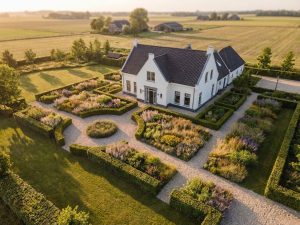 Wit Nederlands landhuis met geometrische tuinen, gravelpad en inheemse beplanting in gouden avondlicht vanuit vogelperspectief